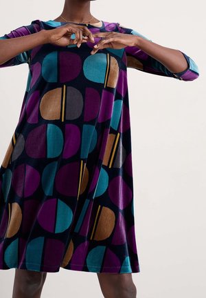 Kobieta nosząca luźną sukienkę z rękawami 3/4, zdobioną geometrycznym wzorem turkusowych, fioletowych, brązowych i czarnych półkoli na czarnym tle.