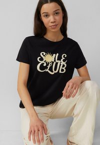 Zwarte katoenen t-shirt met een grote roomkleurige "SOLE CLUB" grafiek met een zonmotief. Model draagt roomkleurige broeken en zit tegen een grijze achtergrond.