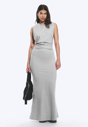 Joven mujer con cabello largo y oscuro lleva un vestido maxi gris ajustado sin mangas y sostiene un bolso negro, de pie contra un fondo claro y liso.