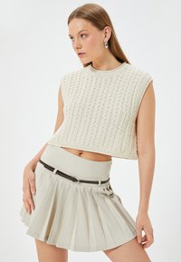 Top corto de punto crema con patrón de trenzado texturizado; combinado con una falda plisada beige claro y un cinturón delgado marrón.