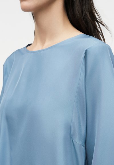 Γυναίκα που φορά ένα απαλό γαλάζιο πουκάμισο με μακριά μανίκια, εμφανίζεται από τον λαιμό μέχρι το πάνω μέρος του στήθους σε απλό φόντο.