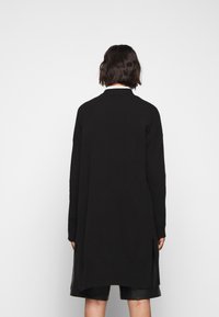 Osoba z krótkimi ciemnymi włosami, ubrana w długi czarny sweter i czarne szorty, stojąca tyłem na tle gładkiego białego tła.