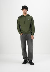 Joven con el cabello rizado corto, vistiendo una sudadera con textura verde, jeans de pierna ancha grises y botas negras, de pie contra un fondo blanco.