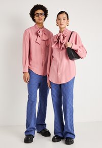 Duas pessoas vestem camisas cor-de-rosa claro combinando, com grandes laços, abotoadas, emparelhadas com calças azuis texturizadas e sapatos pretos robustos.