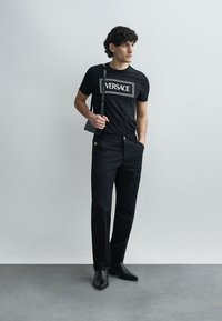 Črna majica s belim napisom "VERSACE", kombinirana s črnimi hlačami in črnimi čevlji. Obleka ima minimalističen dizajn in krojen kroj.