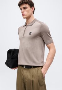 Άνδρας που φοράει πόλο μπλουζάκι πλεκτό μπουφάν με κοντά μανίκια σε χρώμα ταφ με φερμουάρ και λογότυπο HUGO, χακί πλισέ παντελόνια, κρατώντας μια μαύρη τσάντα στο αριστερό χέρι.