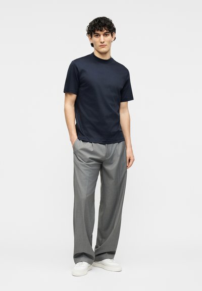 Άνδρας που στέκεται με τα χέρια στις τσέπες, φοράει μπλε κοντομάνικο πουκάμισο, γκρι παντελόνι με φαρδιά πόδια και λευκά αθλητικά παπούτσια σε απλό φόντο.