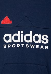Sudadera azul marino hecha de algodón, con un gran logo blanco de "adidas SPORTSWEAR" y un logo rojo del trébol de adidas en la parte superior izquierda.
