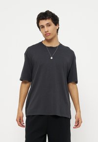 Νέος άνδρας με κοντά σγουρά μαλλιά φοράει μαύρο φαρδύ μπλουζάκι, μαύρο παντελόνι και ασημένιο κολιέ με μενταγιόν μπροστά σε απλό λευκό φόντο.