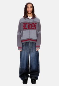 Άτομο με μακριές dreadlocks που φορά γκρι και κόκκινο φούτερ με φερμουάρ και κείμενο "LBN" και φαρδιά φθαρμένα μπλε τζιν με μαύρα παπούτσια.