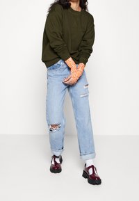 Sudadera verde oliva con puños acanalados, vaqueros azules claros rotos y zapatos burdeos de charol. Mangas de brazo con estampado naranja visibles.