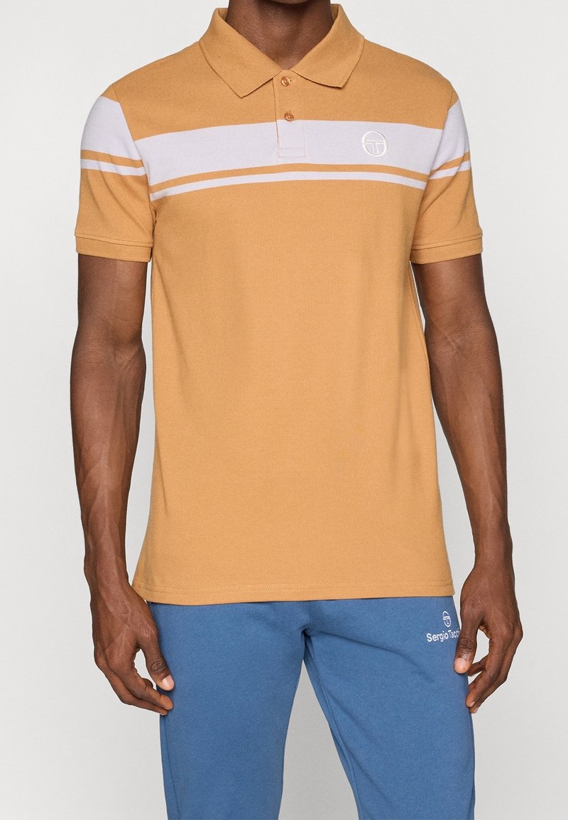 Musztardowa koszulka polo z białymi poziomymi paskami, zapinana na dwa guziki, z małą naszywką logo na piersi. Połączona z niebieskimi spodniami dresowymi.