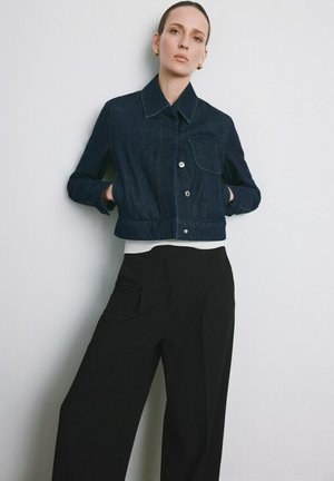 Femme portant une veste en jean courte bleu foncé, un haut blanc, un pantalon large noir, debout les mains dans les poches de la veste, sur fond blanc.