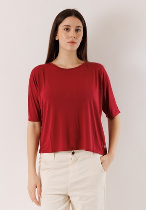 Donna che indossa una maglietta rossa a maniche corte ampia e pantaloni color avorio, con una mano in tasca, in piedi davanti a uno sfondo chiaro uniforme.