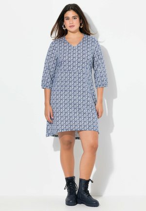 Blau gemustertes Kleid mit V-Ausschnitt, Dreiviertelärmeln und lockerer Passform. Kombiniert mit schwarzen Schnürkampfstiefeln.