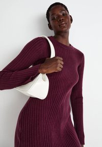 Abito a coste bordeaux con maniche lunghe, caratterizzato da un collo rotondo. Modella che tiene una piccola borsa bianca con design curvo.