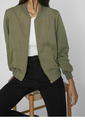 Kvinde iført olivengrøn bomberjakke over hvid top og sorte bukser, siddende på en træskammel mod en grå baggrund.