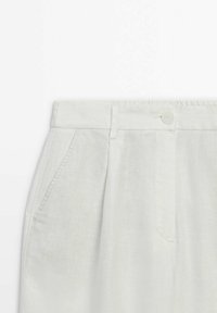 Pantalons en lin beige clair avec fermeture à bouton, devant plissé et poches latérales. Texture lisse et coupe décontractée avec un design épuré.