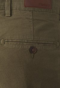 Olivgröna byxor i strukturerad tyg, med en framficka med en brun knapp och en strukturerad brun läderpatch i midjan.
