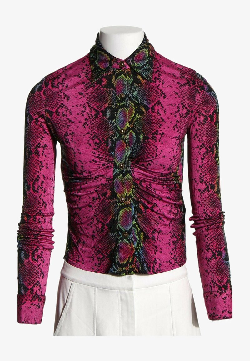 Chemise boutonnée à manches longues avec motif serpent rose et multicolore, détail froncé à l'avant, portée avec un pantalon blanc.