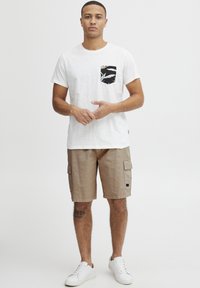 Hvid bomuldst-shirt med en sort lomme med tropisk print, kombineret med beige cargoshorts med sidelommer og hvide sneakers.