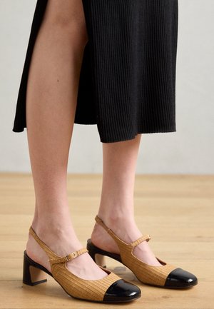 Noszenie beżowych plecionych szpilek typu slingback z czarnymi nakładkami na palce oraz czarnej prążkowanej spódnicy midi na jasnej drewnianej podłodze.
