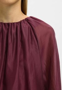 Viininpunainen, kevyt kangastoppi, jossa on rypytetty pääntie ja laskeutuva siluetti, joka korostaa pehmeää tekstuuria ja hillittyä kiiltoa.