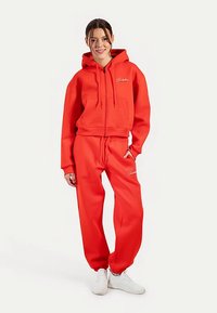 Rode sweatvest met capuchon, voorzien van een voorrits en zakken, gecombineerd met bijpassende trainingsbroek. Heeft een zachte textuur en wit logodetail.