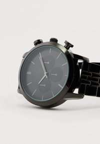 Crni ručni sat s metalnom narukvicom, okruglim brojčanikom, tri kronografska brojčanika i srebrnim oznakama sati na jednostavnoj svijetloj pozadini.