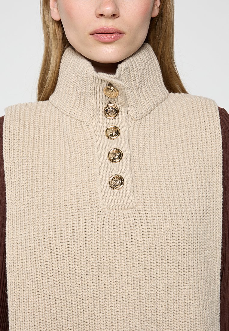 Nahaufnahme einer Frau in einem beigen, gestrickten ärmellosen Pullover mit hohem Kragen und fünf goldenen Knöpfen auf der Vorderseite.