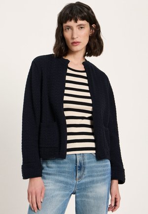 Frau mit kurzen dunklen Haaren, die eine schwarze, strukturierte, offene Strickjacke, ein schwarz-weiß gestreiftes Oberteil und hellblaue Jeans vor schlichtem Hintergrund trägt.