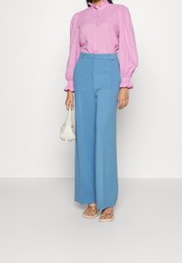 Light pink textured blouse with ruffled collar, paired with high-waisted, wide-leg blue trousers. Holding a small white handbag, wearing clear sandals.