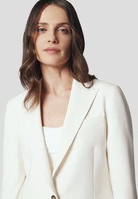 Biała żakardowa marynarka o gładkim materiale z klapami o wyznaczonych rogach, z jedynym zapięciem na guzik i kieszenią na piersi; noszona na białej bluzce.