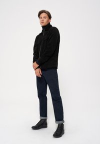 Czarna kurtka sztruksowa na zamek, z dwiema kieszeniami na piersi; połączona z ciemnoniebieskimi spodniami dżinsowymi z podwiniętymi nogawkami oraz czarnymi butami na sznurówki. Prosty design i gładka tekstura.