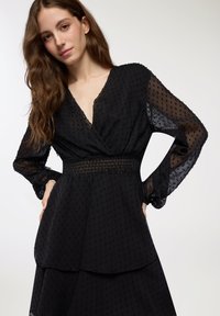 Abito nero realizzato in tessuto trasparente e testurizzato con motivo a pois. Presenta una profonda scollatura a V, vita arricciata e maniche lunghe e fluide.