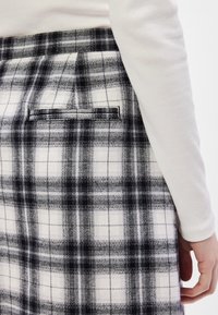 Μαύρη και λευκή καρό φούστα με υφαντή υφή, διαθέτοντας πτυχώσεις και πίσω τσέπη. Στενή σχεδίαση με ένα λείο, λευκό μπλουζάκι μακρυμάνικο που διακρίνεται μερικώς.