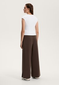 Žena stojaca obrátene vľavo, oblečená v bielom bezrukávovom topu, širokých hnedých nohaviciach a bielych teniskách na svetlom pozadí.