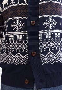 Cardigan tricotat de marina, cu motive Fair Isle în alb și maro, manșete ribbate și nasturi maronii de-a lungul plăcii frontale.