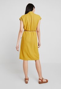 Hořčicově žluté lněné šaty s krátkými rukávy ve stylu cap, zúženým pasem pomocí stahovací šňůrky a délky do kolen. Nošeny s hnědými sandály.