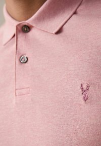 Polo de color rosa claro, hecho de tejido texturizado, que presenta un cierre de dos botones y un pequeño logo bordado de un ciervo en el lado izquierdo del pecho.