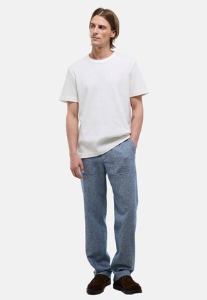 Homme aux cheveux mi-longs portant une chemise blanche unie à manches courtes, un pantalon bleu avec des poches avant et des chaussures brun foncé, debout, détendu.