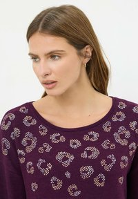 Maglione viola con scollatura ampia, caratterizzato da un motivo leopardato di borchie dorate e argentate sulla parte anteriore. Tessuto morbido, design casual.