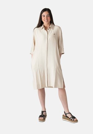 Mujer sonriente con cabello largo y oscuro, vistiendo un vestido camisa beige con rayas hasta la rodilla y sandalias de plataforma negras, de pie con las manos en los bolsillos.