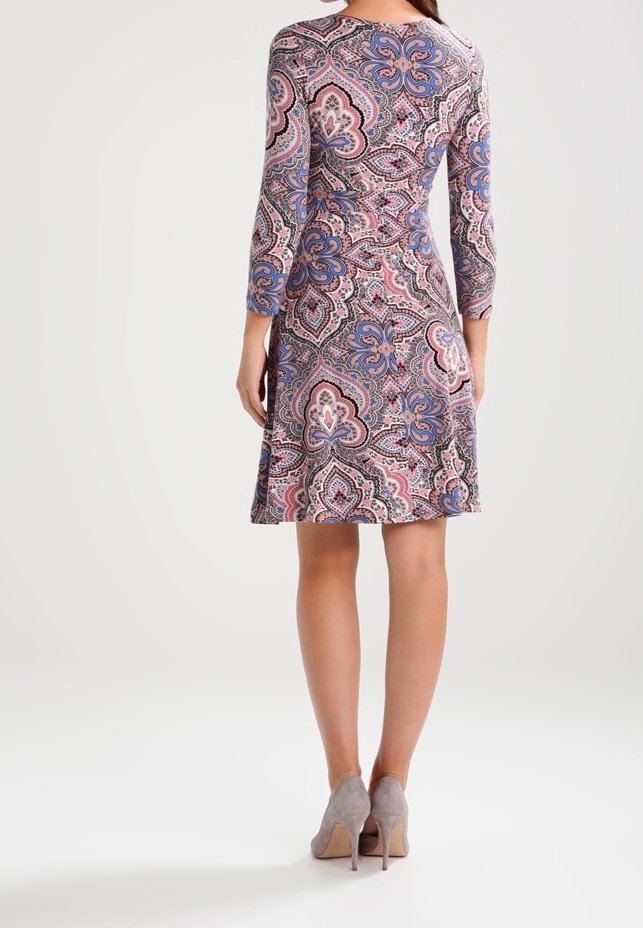 Femme portant une robe longue jusqu'aux genoux et à manches longues avec des motifs floraux roses, bleus et noirs, associée à des chaussures à talons gris, vue de dos.