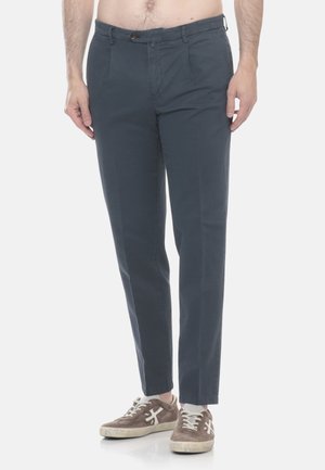 Uomo che indossa pantaloni slim fit grigio scuro e sneaker in camoscio marrone con lacci bianchi, in piedi su sfondo bianco.