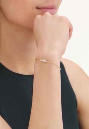 Vrouw draagt een fijn gouden armbandje met een hartvormige bedel, rust haar vuist licht tegen haar kin, draagt een mouwloze zwarte top.