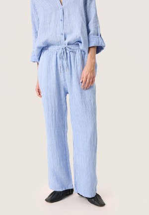 Femme portant un pantalon en lin à rayures verticales bleues et blanches, léger et ample, avec une taille à cordon de serrage, et une chemise à boutons assortie, ainsi que des chaussures plates noires.