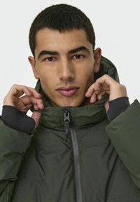 Chaqueta aislante verde con un cuello alto y cremallera. Presenta detalles en negro en los puños, ofreciendo un diseño y textura contemporáneos.