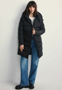 Czarna kurtka puchowa z kapturem, wyposażona w zamek z przodu i kieszenie. Połączona z jasnoniebieskimi spodniami o szerokich nogawkach i czarnymi butami.