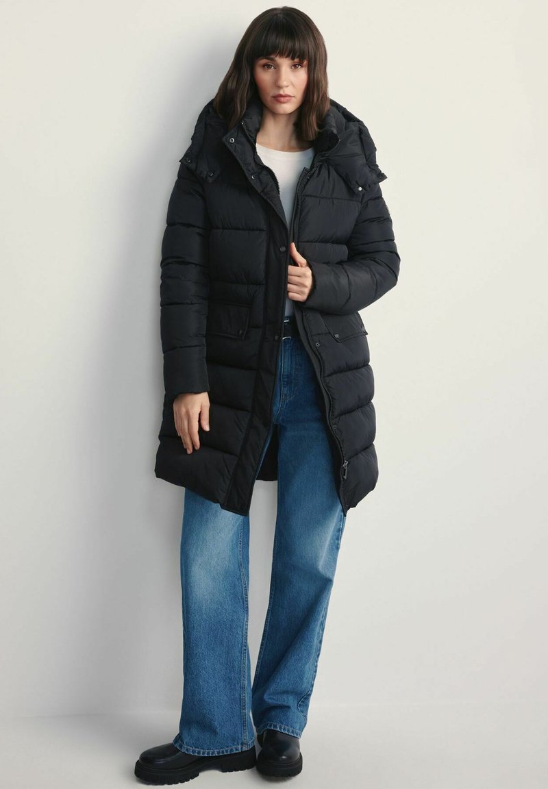 Czarna kurtka puchowa z kapturem, wyposażona w zamek z przodu i kieszenie. Połączona z jasnoniebieskimi spodniami o szerokich nogawkach i czarnymi butami.